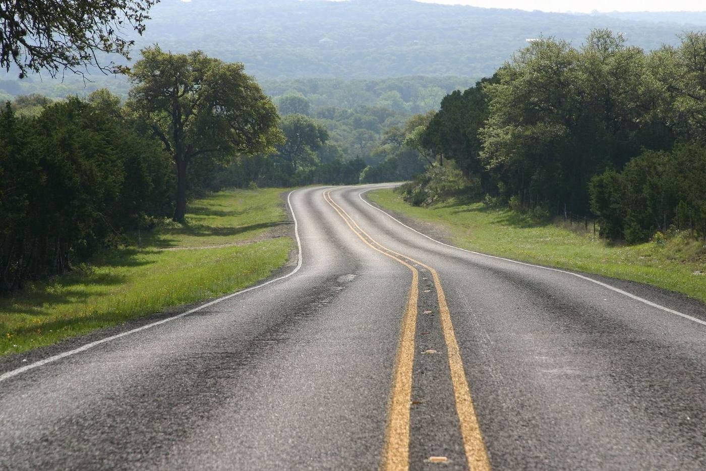 A two-lane ranch road curving through live oaks in the Texas Hill Country